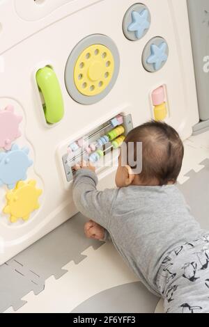 Six month old baby boy on stomach in playpen, reaching to spin beads on wire on busy box toy Stock Photo