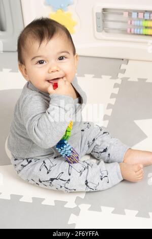 Six month old baby boy sitting on floor smiling portrait looking at camera holding hand with small toy near mouth Stock Photo