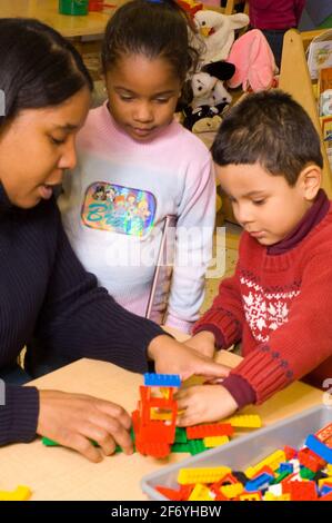 Preschool classroom 3-4 year olds female teacher talking with boy about the structure he has made with Lego plastic bricks as girl holding book looks Stock Photo