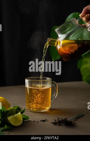 Woman pouring hot black tea with mint, lemon and lime from teapot into glass mug, evening tea party Stock Photo