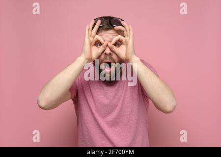 Bizarre, shocked man put fingers to eyes like binoculars and looks through them with mouth open in surprise. Pink background. Stock Photo
