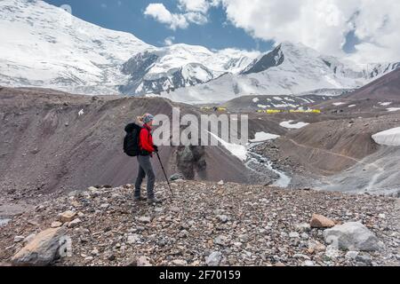 Young woman with a large backpack and trekking poles looking at the base camp of snowcapped mountain with yellow camping tents in front of huge massif Stock Photo