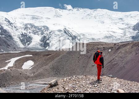 Man with a large red backpack examining the climbing route up to the summit of huge snowcapped mountain Lenin Peak on approach to base camp in Pamir Stock Photo