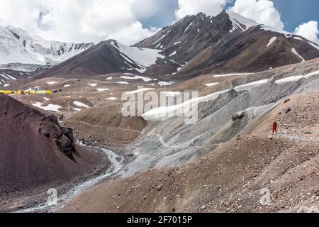 Adult man with a large backpack approaching the base camp in Pamir mountain range with enormous snowcapped Lenin Peak in distance, Kyrgyzstan Stock Photo