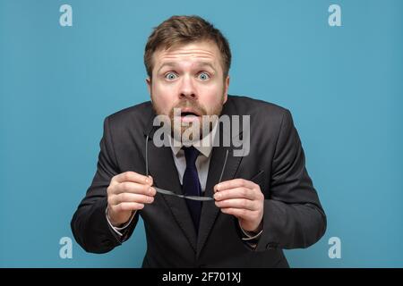 Shocked, frightened business man in a suit has taken off glasses and looks with big eyes opening mouth. Blue background. Stock Photo
