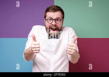 Bizarre bearded man in round glasses makes an approving gesture with hands thumbs up and joyfully looks into the camera.  Stock Photo