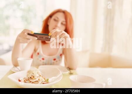 Red-haired girl takes photos of salad on smartphone camera. Woman at table on terrace in cafe. Mobile phone in brown case. White ceramic dishes on table. Salad of chicken, eggs and cheese. Stock Photo