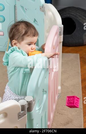 10 month old baby girl standing with support throwing block over playpen barrier Stock Photo