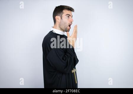 Young hispanic man wearing priest uniform standing over white background covering mouth with hand, shocked and afraid for mistake. surprised expressio Stock Photo