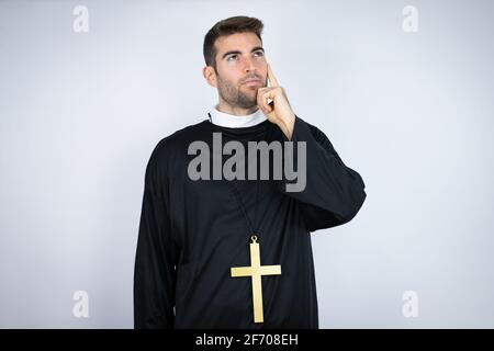 Young hispanic man wearing priest uniform standing over white background serious face thinking about question with hand on chin, thoughtful about conf Stock Photo