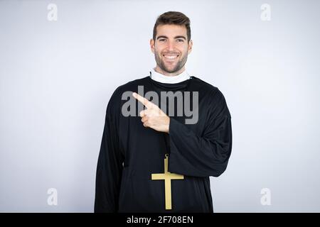 Young hispanic man wearing priest uniform standing over white background smiling and pointing with hand and finger to the side Stock Photo