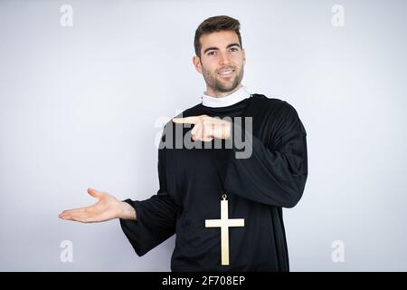 Young hispanic man wearing priest uniform standing over white background surprised, showing and pointing something that is on his hand Stock Photo