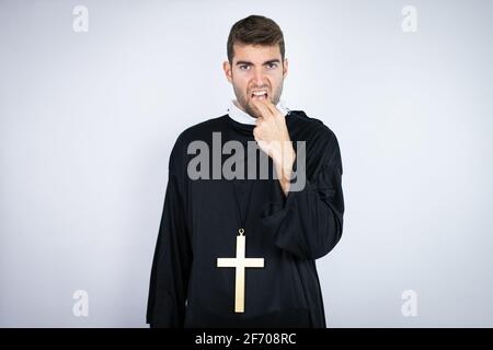 Young hispanic man wearing priest uniform standing over white background disgusted with his hand inside his mouth Stock Photo