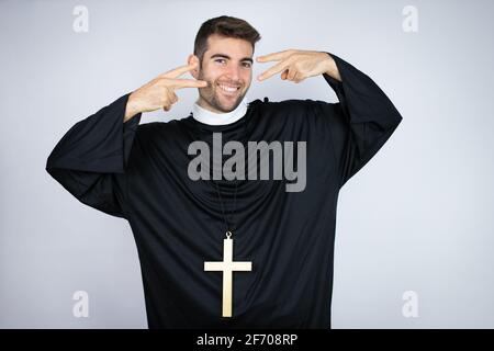 Young hispanic man wearing priest uniform standing over white background Doing peace symbol with fingers over face, smiling cheerful showing victory Stock Photo