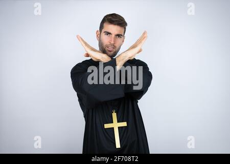 Young hispanic man wearing priest uniform standing over white background Rejection expression crossing arms doing negative sign, angry face Stock Photo