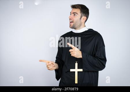 Young hispanic man wearing priest uniform standing over white background surprised and pointing her fingers side Stock Photo