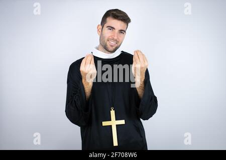 Young hispanic man wearing priest uniform standing over white background doing money gesture with hands, asking for salary payment, millionaire busine Stock Photo