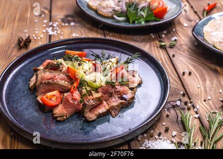 Beef tenderloin fried on fire with herbs, seasoned with spices, tomatoes. Wooden brown background decorated with rosemary, spices. Restaurant menu. Stock Photo