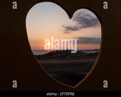 View through the fence hole in the shape of heart. Man sitting thougtfully at the seashore and watching orange sun setting down. Love, care and lonely Stock Photo