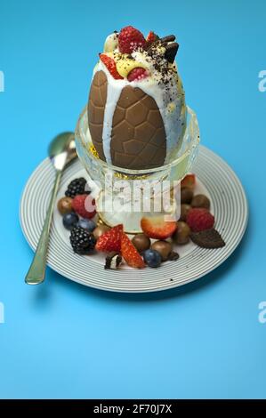 Beautiful vertical view of delicious chocolate Easter egg topped with ice cream, berries, cookies, custard with spoon on light blue background Stock Photo
