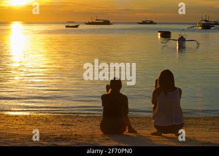 Silhouetted shape of two friends sitting on the beach at the shore of the Visayan sea at sunset. Logon village, Malapascua Island.  Cebu, Philippines Stock Photo