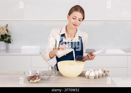 Female confectioner in blue apron pouring milk in in big bowl. Process of making a pie or muffins Stock Photo