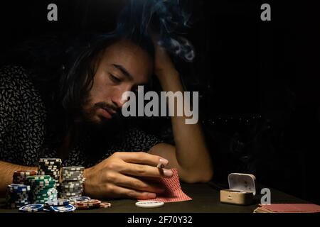 man playing poker while looking at cards and smoking tobacco. young long-haired man, gambling, dramatic light. Stock Photo