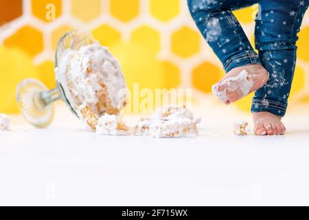 A small child's feet smeared in cream, and a cake lying on the floor in close-up. In the background is a pattern of yellow honeycombs and balloons. Co Stock Photo