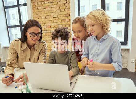 Curious learners. Young female teacher in glasses showing scientific robotics video to joyful children using laptop during STEM class Stock Photo