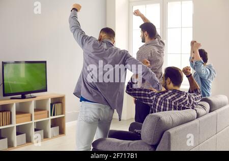 Active adult male friends with arms raised emotionally watching a football match on TV at home. Stock Photo