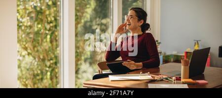 Smiling woman designer at home. Female graphic designer sitting at table with digital tablet looking away and smiling. Stock Photo