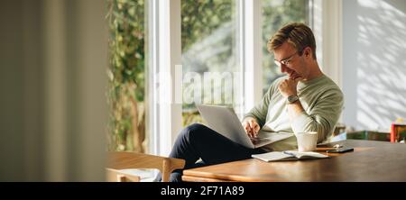 Young man using laptop and smiling at home. Man sitting by table working on laptop computer. Stock Photo