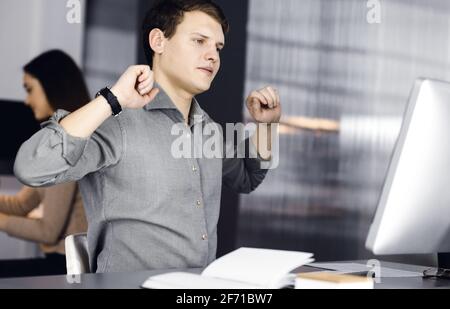 Young businessman and programmer with brown curly hair in a green shirt is stretching his hands after working hard on computer, sitting together with Stock Photo