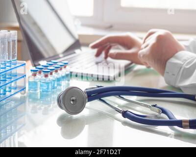 Vaccination medical research concept. Focus on the stethoscope. Hands of a doctor working at a laptop. He fills out the patient's questionnaire online Stock Photo