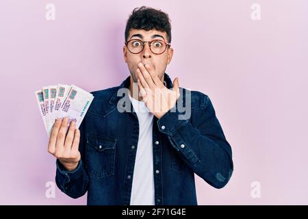 Young hispanic man holding egyptian pounds banknotes covering mouth with hand, shocked and afraid for mistake. surprised expression Stock Photo