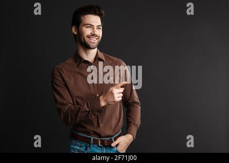 Smiling handsome unshaven guy pointing finger aside isolated over black background Stock Photo