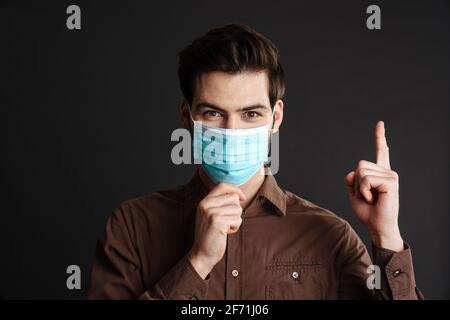 Surprised man posing in protective mask and pointing finger upward isolated over black background Stock Photo