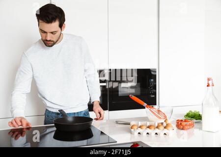Focused handsome young man cooking scrambled eggs at home kitchen Stock Photo