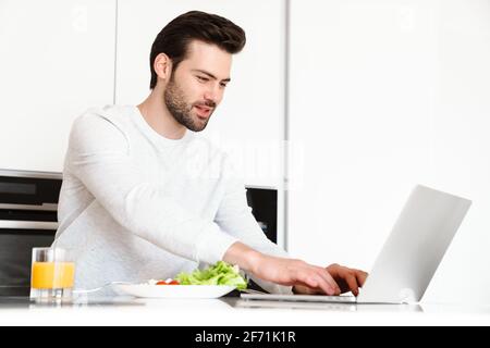 Pleased handsome man using laptop while having breakfast at home kitchen Stock Photo