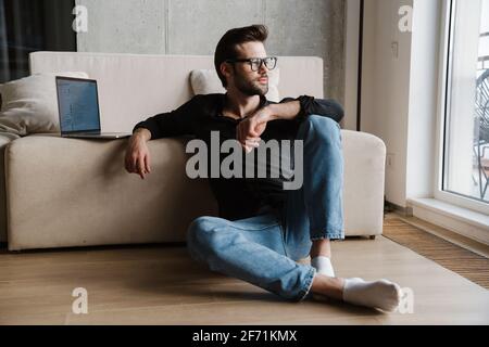 Pleased unshaven man using laptop while sitting on floor at home Stock Photo