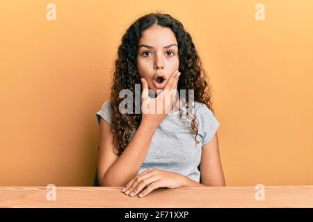 Teenager hispanic girl wearing casual clothes sitting on the table looking fascinated with disbelief, surprise and amazed expression with hands on chi Stock Photo