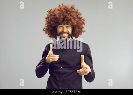 Funny man in crazy big curly wig pointing at camera standing on gray studio background Stock Photo
