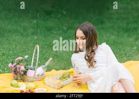 A beautiful young woman lies on a yellow blanket on the grass, holding a glass of wine Stock Photo