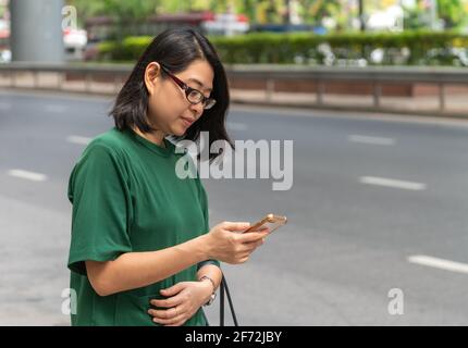 Asian middle-aged woman looking at smartphone in hand, wearing eyeglasses, standing on street sidewalk. Concept for waiting and using smartphone to tr Stock Photo