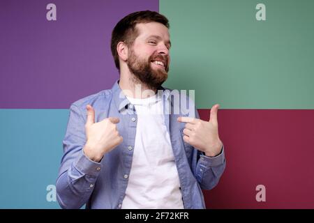 Attractive smiling man points his fingers at himself and looks pleased. Colorful background. Stock Photo