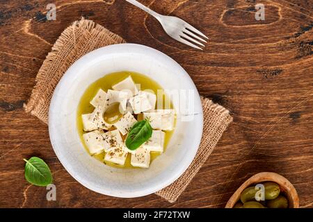 Feta cheese cubes with rosemary, olives and olive oil sauce in white bowl on old brown wooden background. Traditional Greek homemade cheese. Selective Stock Photo