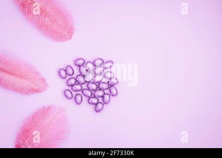 Easter pink background with chocolate easter eggs wrapped in purple colored foil with colorful feathers on a light rose background. Easter concept. Aesthetic creative still life composition.  Stock Photo