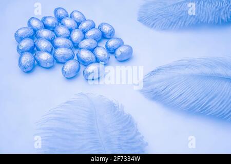 Easter blue light background with chocolate easter eggs wrapped in foil with blue feathers on a light blue background. Easter concept. Aesthetic creat Stock Photo