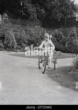 1956, historical, outside in an enclosed area, on a paved path, a young boy in shorts and sandels, riding a tricycle, England, UK. Stock Photo