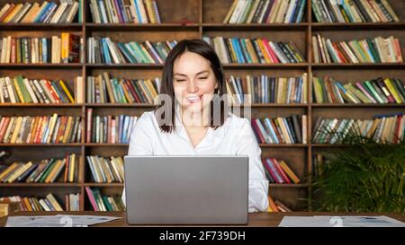 Positive lady in stylish white blouse takes part in online conference via contemporary laptop at table against racks full of books in office Stock Photo